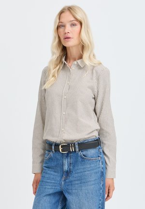 Femme blonde portant une chemise beige et blanche à rayures boutonnée rentrée dans un jean bleu avec une ceinture noire, debout devant un fond uni.