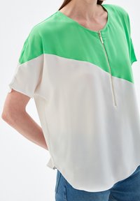 Blouse à manches courtes avec une partie supérieure verte et une partie inférieure blanche, dotée d'une fermeture éclair frontale et d'un tissu lisse et léger.