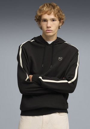 Junge Person mit kurzen, lockigen blonden Haaren, trägt einen schwarzen Hoodie mit weißen Streifen und beige Hose, die Arme verschränkt, neutraler Gesichtsausdruck.