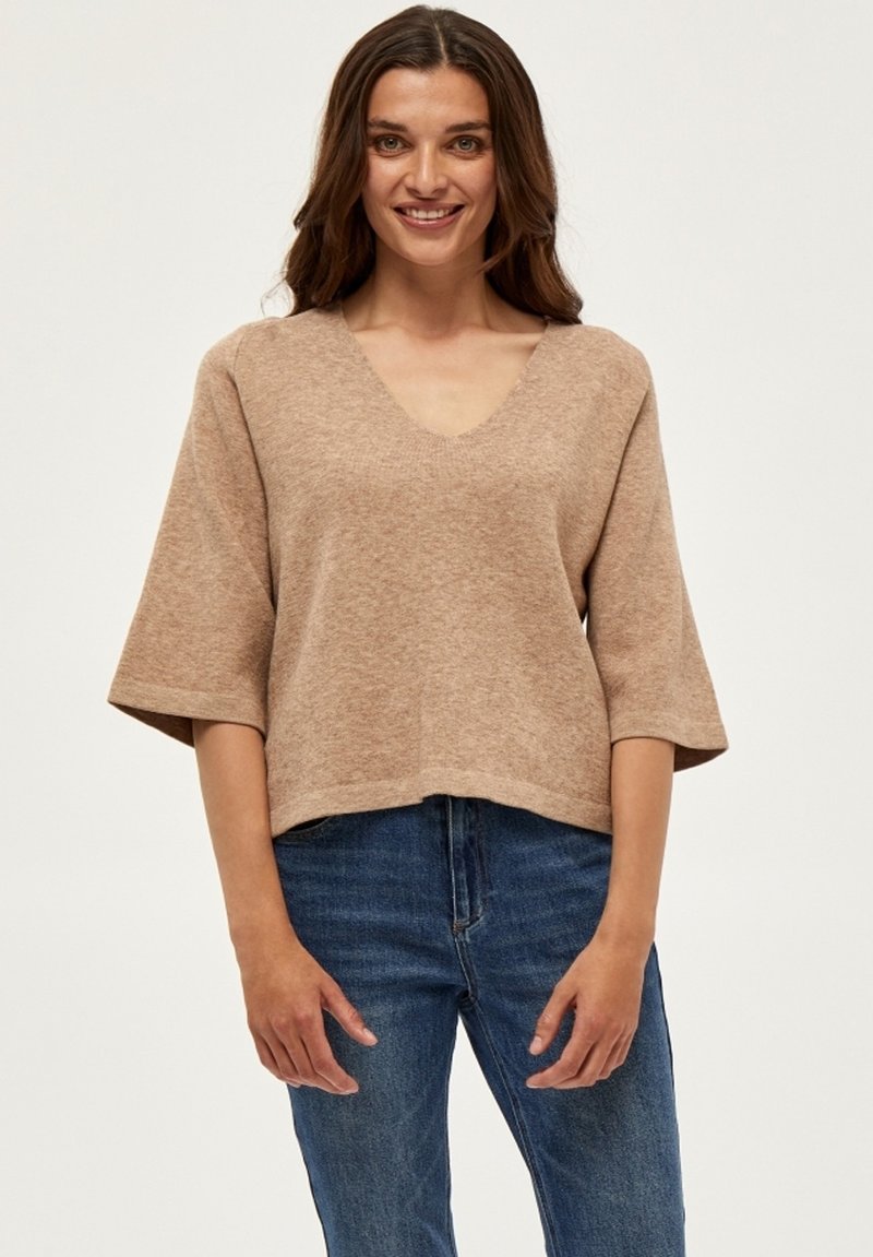 Beige strikket sweater med V-hals, brede korte ærmer og en afslappet pasform, parret med blå denim jeans. Tekstureret, blødt materiale.