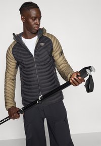 Μπουφάν με επένδυση σε μαύρα και μπεζ τμήματα, με φερμουάρ στο μπροστινό μέρος, λογότυπο στο στήθος και κουκούλα, κρατημένο με ένα μπαστούνι πεζοπορίας.