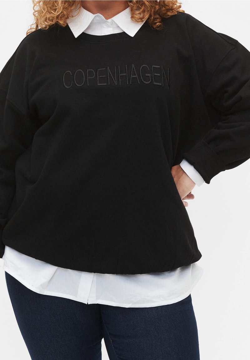 Persona con sudadera negra con el texto "COPENHAGEN", con una camisa blanca de cuello debajo y jeans azules, mano en la cadera.