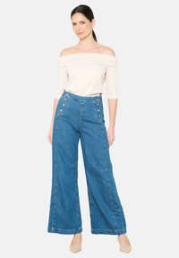 Gaiši bežs off-the-shoulder tops un augsto jostasvietu zili džinsu platā kāju bikses ar pogu akcentiem priekšpusē. Gluda tekstūra, strukturēts dizains.