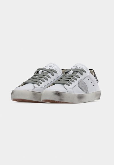 Paire de baskets blanches basses avec des lacets gris, un patch logo gris, et des semelles légèrement sales sur un fond blanc.