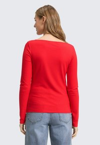 Rotes geripptes Langarmshirt mit weitem Ausschnitt. Der Stoff hat eine glatte Textur und wird mit hochgeschnittenen hellblauen Jeans kombiniert.