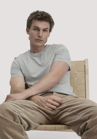 Jonge man met krullend haar, gekleed in een lichtgrijze t-shirt en beige corduroy broek, zittend op een geweven stoel en recht vooruit kijkend.