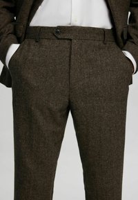 Barna tweed nadrág texturált felülettel, ferde zsebekkel és egy gombos zárással a deréknál.