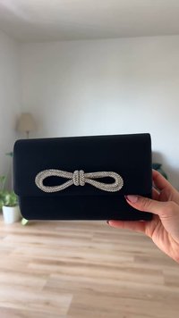 Čierna kabelka na clutch s textúrovaným povrchom, s ozdobným uzáverom vo forme kamienkového mašľového zapínania. Kompaktný obdélnikový tvar, držaná v ruke.