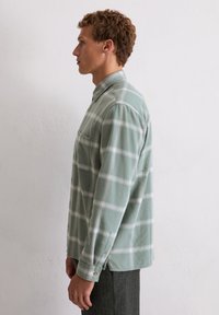 Jonge man in een groen-wit geruite lange mouwen shirt, die zijwaarts tegen een effen witte textuurmuur staat.