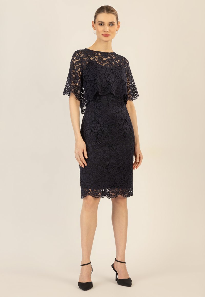 Marineblaues Spitzenkleid mit einer mehrschichtigen Umhangüberlagerung, die Blumenmuster und einen gewellten Saum aufweist. Glatte Textur, knielänge Passform, kombiniert mit schwarzen Absätzen.