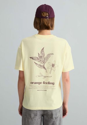 Osoba nosząca bladoróżowy t-shirt z botanicznymi rysunkami liniowymi i tekstem "orange feeling" na plecach, połączony z wyblakłymi szarymi jeansami i fioletową czapką.