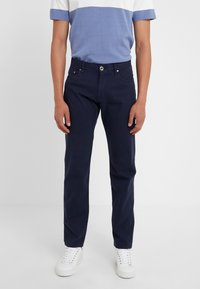 Námořnické modré bavlněné kalhoty s rovnými nohavicemi, které mají pět kapes a zapínání na předním zipu, nošené s šedou a modrou pruhovanou košilí.