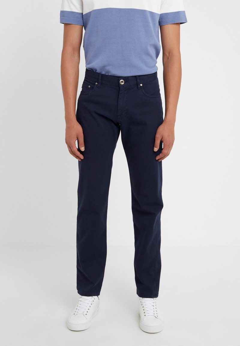 Námořnické modré bavlněné kalhoty s rovnými nohavicemi, které mají pět kapes a zapínání na předním zipu, nošené s šedou a modrou pruhovanou košilí.