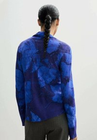 Lila Langarmhemd mit blauem Blumenmuster, glattem Stoff, lockerer Passform und abgerundetem Saum, getragen mit grauen strukturierten Hosen.