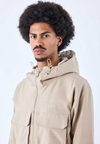 Chaqueta con capucha beige de acabado texturizado, que cuenta con dos bolsillos frontales y un logotipo bordado de "Project x Paris". Cuerdas de ajuste en la capucha.