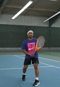 Τενίστας με λευκό περιουσάς, φορώντας μωβ μπλούζα με σχέδιο και μαύρα σορτς, κρατώντας μια πορτοκαλί ρακέτα τένις σε γήπεδο.