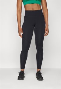 Fekete leggings magas derékkal, ívelt varratokkal és rugalmas anyagból, fekete sportcipővel és zöld crop top-ról van szó.