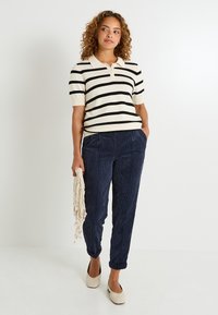 Polo de manga corta a rayas en crema y negro, combinado con pantalones de pana en azul marino. El modelo sostiene un bolso con flecos y lleva zapatos de color claro.