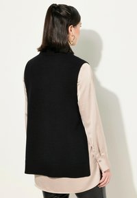 Gilet senza maniche nero a maglia con orlo a coste, indossato sopra una camicia a maniche lunghe di colore chiaro e in raso. Texture liscia, vestibilità casual.