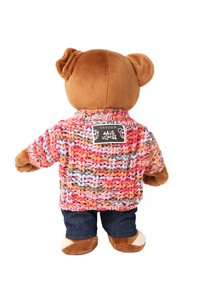 Nallebjörn som bär en flerfärgad stickad tröja med rosa, blå och bruna trådar samt blå jeans. En etikett på tröjans baksida är synlig.