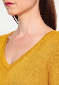 Femme aux longs cheveux bruns portant un pull en maille en V couleur moutarde, dévoilant son cou, son épaule et le bas de son visage.