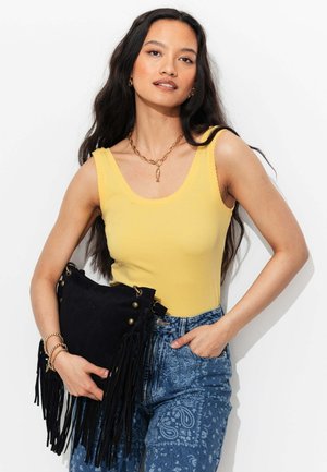 Femme aux longs cheveux foncés portant un haut jaune sans manches et un jean bleu à motifs, tenant un sac à main noir à franges sur fond blanc.