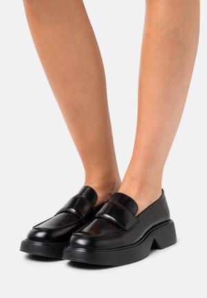 Mocassins en cuir noir avec une large sangle, un embout carré et une semelle épaisse noire. Texture lisse avec des accents minimaux.