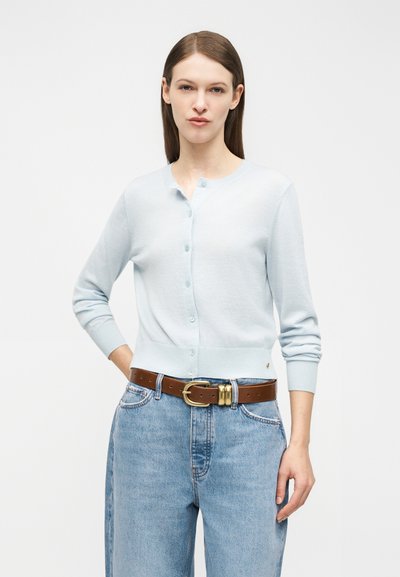 Kvinne med rett brunt hår iført en lyseblå cardigan med knapper, høytlivs lyseblå jeans og et brunt belte med messingspenne.