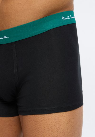 Κοντινό πλάνο ενός ατόμου που φοράει μαύρα μπόξερ με πράσινη μέση που φέρει το λογότυπο "Paul Smith" σε λευκό χρώμα.