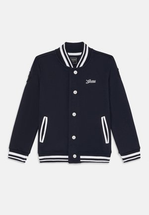 Veste varsity bleu marine avec col, poignets et poches à rayures blanches, boutons sur le devant et logo Guess sur la poitrine gauche.