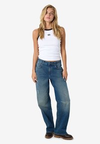 Witte geribde tanktop met zwarte accenten, gecombineerd met loszittende blauwe spijkerbroek met vervaagde delen en voorzakken. Bruine laarzen maken de look af.