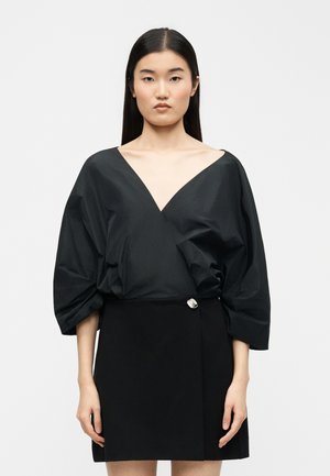 Hosszú sötét hajú nő fekete, mély V-nyakú felsőt visel, amelynek bő ujjai vannak, valamint egy fekete szoknyát ezüst gombdísszel.