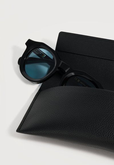 Lunettes de soleil à monture noire avec verres bleu foncé partiellement à l'intérieur d'un étui en cuir noir texturé sur une surface blanche.