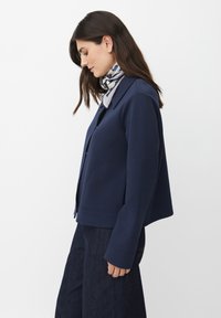 Kobieta z ciemnymi włosami, ubrana w granatową kurtkę, ciemne dżinsy i wzorzysty szalik, stojąca w profilu na białym tle.