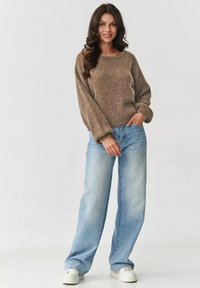 Brązowy sweter dzianinowy o luźnym kroju i szerokich rękawach, w połączeniu z jasnoniebieskimi spodniami typu wide-leg i białymi sneakersami na jednolitym tle.