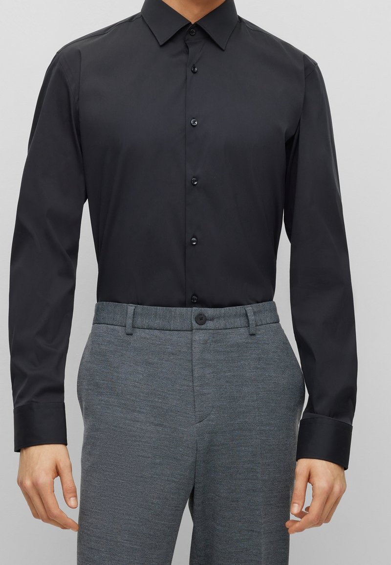 Homme portant une chemise noire à manches longues boutonnée rentrée dans un pantalon de costume gris sur un fond uni.