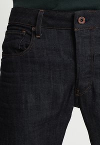 Tmavě modré džíny z denimu s klasickým pětikapsovým designem, měděným kováním, oranžovým šitím a střihem s rovnými nohavicemi.