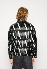 Giacca a vento nera con finitura lucida, caratterizzata da ampie strisce diagonali bianche e un colletto alto. Spessa imbottitura con cuciture visibili.