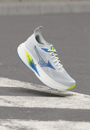Scarpa da corsa atletica bianca e blu con accenti giallo neon, sospesa sopra l'asfalto e sollevando piccole particelle di sporco.