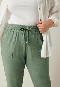 Groene linnen broek met een trekkoord in de taille, voorzien van voorzakken en een gestructureerde stof. Gepaard met een wit shirt.