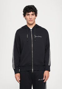 Black zip-up hoodie with white stripes along the sleeves and logo on the chest. Made from soft fabric, featuring a drawstring hood and ribbed cuffs.