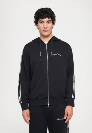Svart zip-up hettegenser med hvite striper langs ermer og logo på brystet. Laget av mykt stoff, med snorhette og ribbede mansjetter.