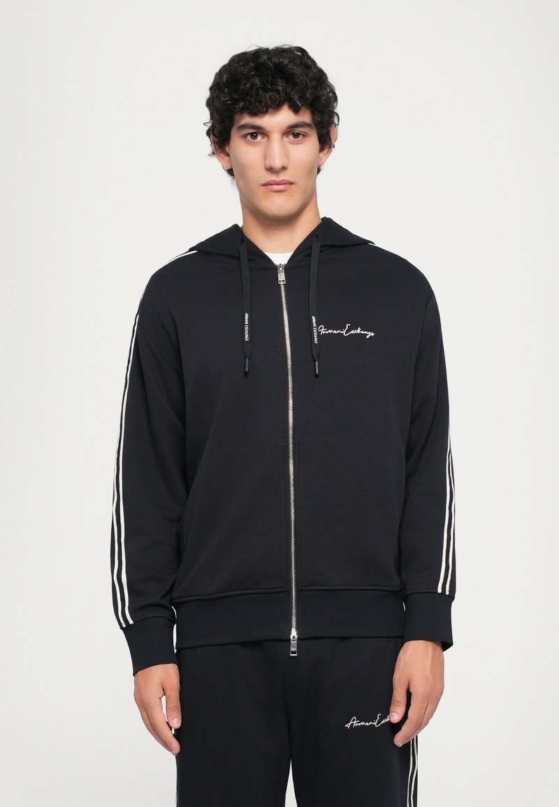 Black zip-up hoodie with white stripes along the sleeves and logo on the chest. Made from soft fabric, featuring a drawstring hood and ribbed cuffs.