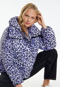 Jachetă pufoasă cu un imprimeu leopard violet și negru, guler mare, fermoar în față și buzunare laterale. Purtați cu pantaloni negri într-o poziție așezată.