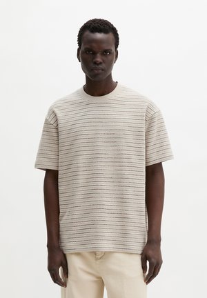 Junger Mann trägt einen beige kurzärmligen Strickpullover mit dünnen horizontalen Streifen und hellfarbene Hosen vor einem schlichten weißen Hintergrund.