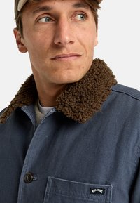 Blue fabric jacket featuring a brown fuzzy collar, button closure, and a chest pocket with a logo tag. The texture appears durable.