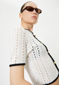 Weiße, gestrickte, cropped Strickjacke mit schwarzem Besatz, kurzen Ärmeln und goldenen Knöpfen. Verfügt über ein strukturiertes, offenes Gewebemuster.