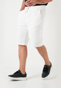 Shorts albi din material elastic, cu talie cu elastic și șnur, având buzunare laterale și o textură netedă și moale. Purtați cu pantofi sport negri.