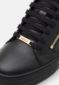 Baskets en cuir noir avec lacets plats noirs, détail doré sur l'étiquette des lacets et semelle en caoutchouc texturée. Dotées d'une fermeture éclair latérale.