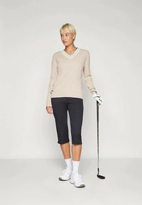 Béžový rebrovaný sveter s výstrihom do V a svetlomodrým lemom, v kombinácii s čiernymi nohavicami po kolená, bielymi ponožkami a golfovými topánkami, v ruke drží golfovú palicu.